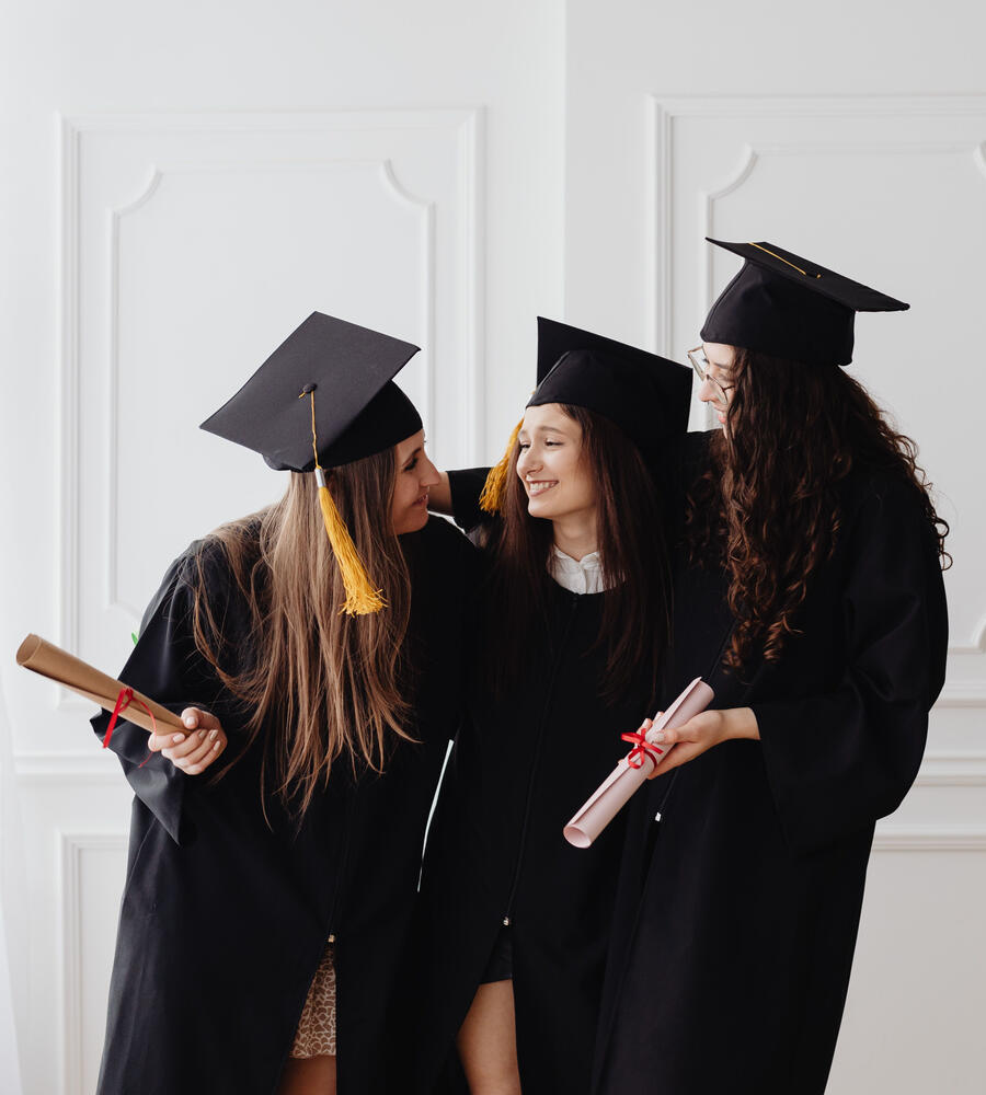 Three graduates in robes standing together and smiling at each other on a while wall background.